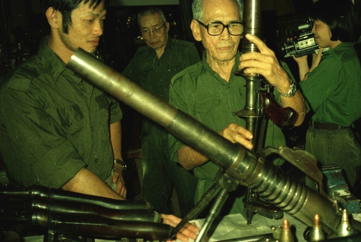 1989年8月7日，日籍军工老战士田中清明（党内化名“清德”，中右戴黑框眼镜手扶炮管者）与桥本惠之（党内化名“阿福”，中左戴眼镜者）在勿洞密林地下兵工厂内同青年军械员检视八九式重掷弹筒与迫击炮管，旁侧女战士正持摄像机做历史记录。