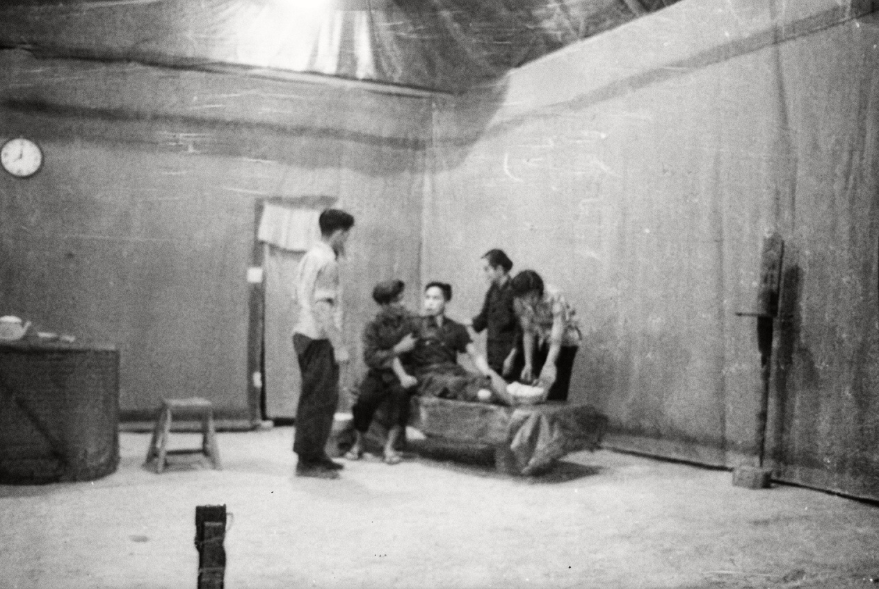 1968年密林营地医疗站场景，医护人员在简易木床旁照料并包扎伤病员。