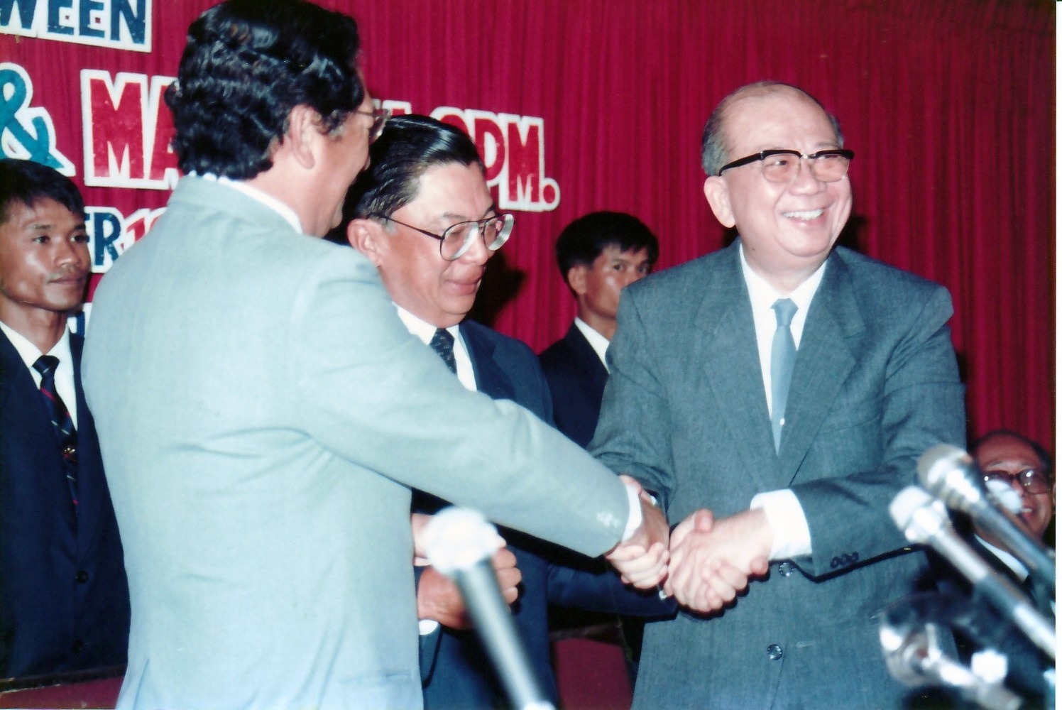 1989年12月2日《合艾和平协议》签署仪式后，马共中央总书记陈平（右）在合艾蠡园酒店主席台上与泰方及马方代表团首席代表微笑握手，标志着和平之门正式敞开。