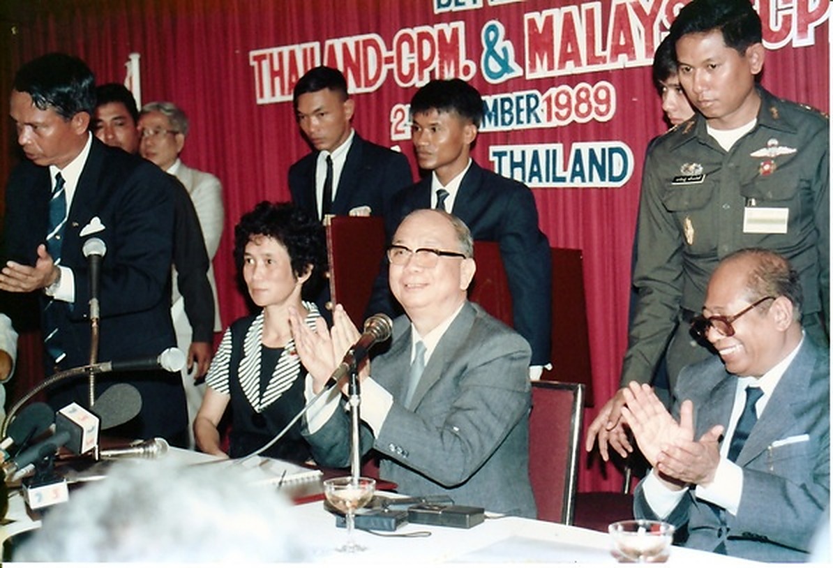 1989年12月2日合艾蠡园酒店签约大典：马共中央总书记陈平（前排中）与党主席阿都拉·西迪（前排右）在主席台就座并鼓掌致意，身后悬挂三方联合签署和平协议的巨幅英文字幕。